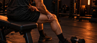 Sportif assis sur un banc de musculation qui masse l'avant de sa cuisse courbaturée, haltère léger posé au sol