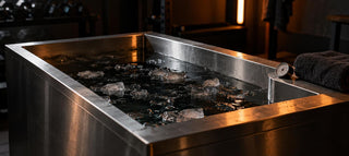 Cuve de bain froid en inox remplie d'eau sombre et de morceaux de glace, thermomètre sur le rebord, dans une salle de sport sombre