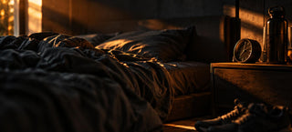 Lit défait dans une chambre sombre au petit matin, réveil, bouteille d'eau et chaussures de sport posés sur la table de nuit